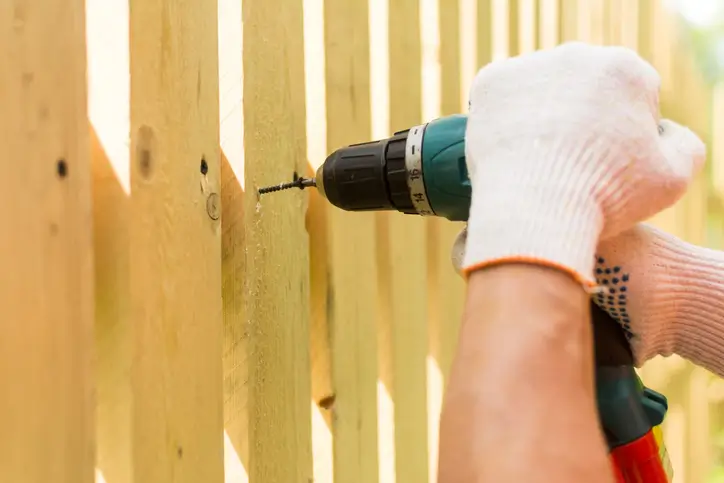 Hands of a carpenter holding an electric screwdriver while repairing a fence